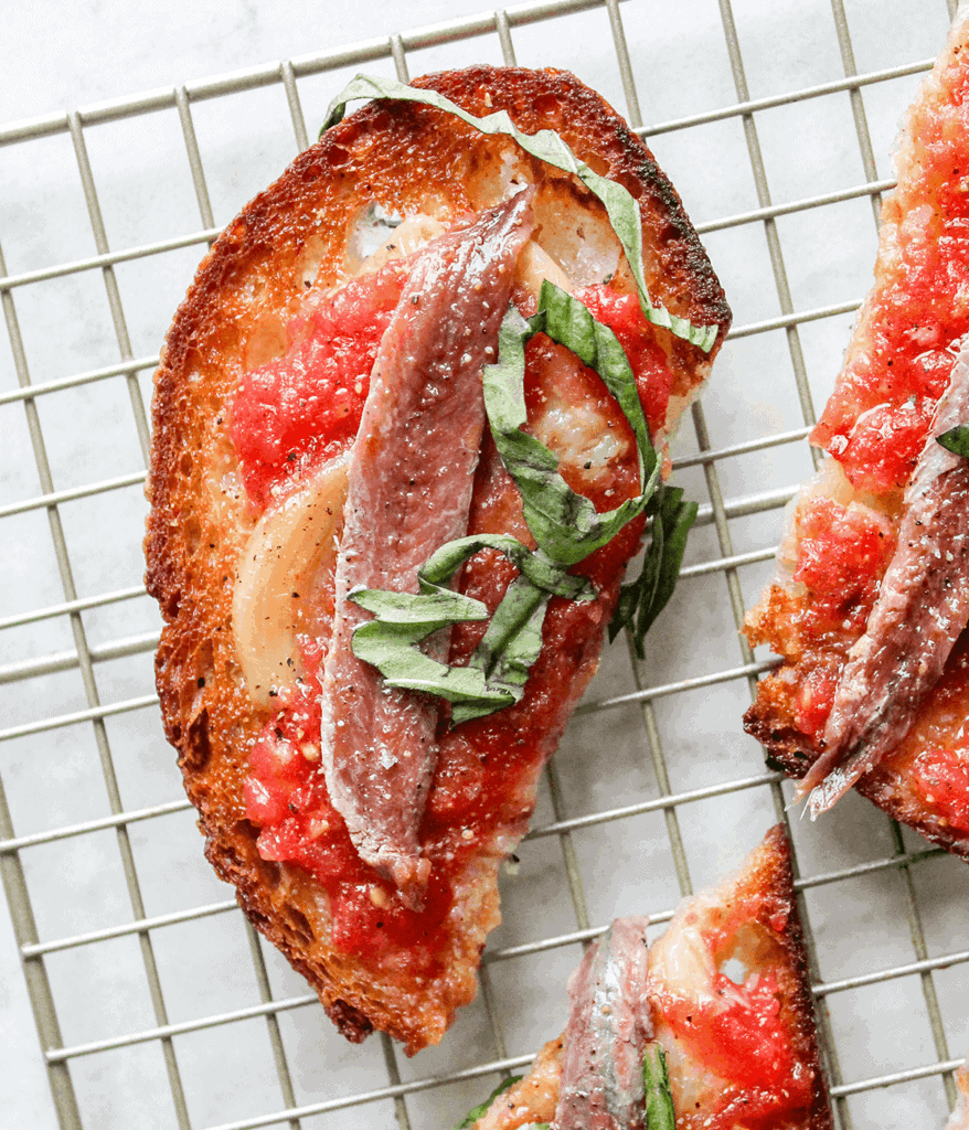 Brightly colored Pan Con Tomate topped with fresh anchovies and chopped herbs, served on a cooling w.