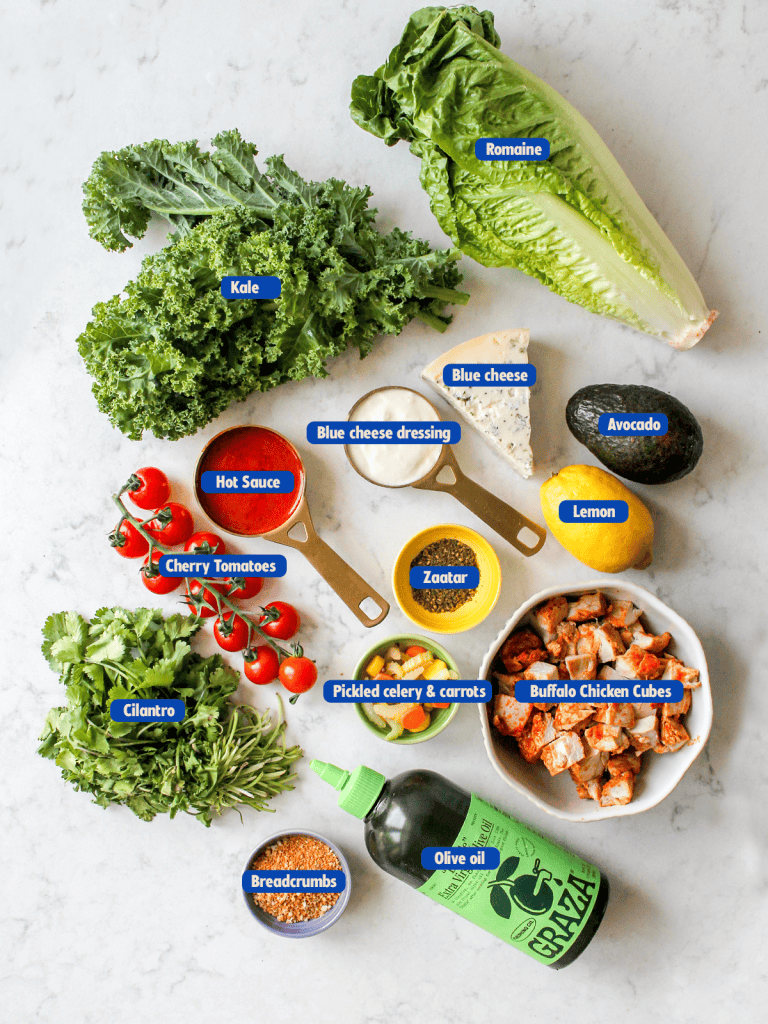 Ingredients for buffalo chicken salad which include kale, romaine, blue cheese, avocado, lemon, hot sauce, zaatar, cherry tomatoes, pickled celery and carrots, buffalo chicken, cilantro, olive oil, breadcrumbs.