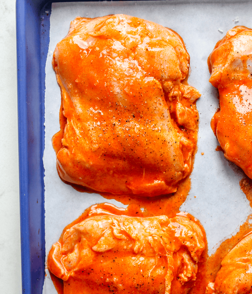 Raw chicken coated in hot sauce on a parchment-lined baking sheet.