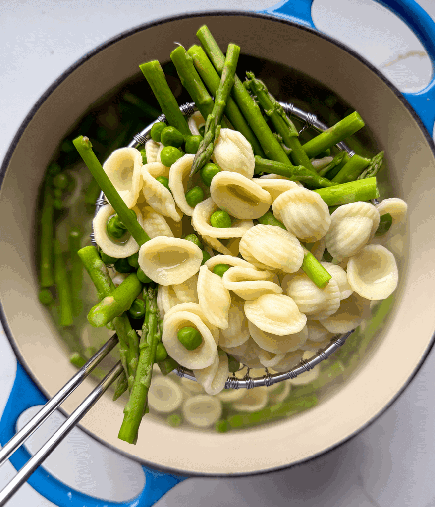 Strainer with pasta, peas, and asparagus with a blue pot underneath.